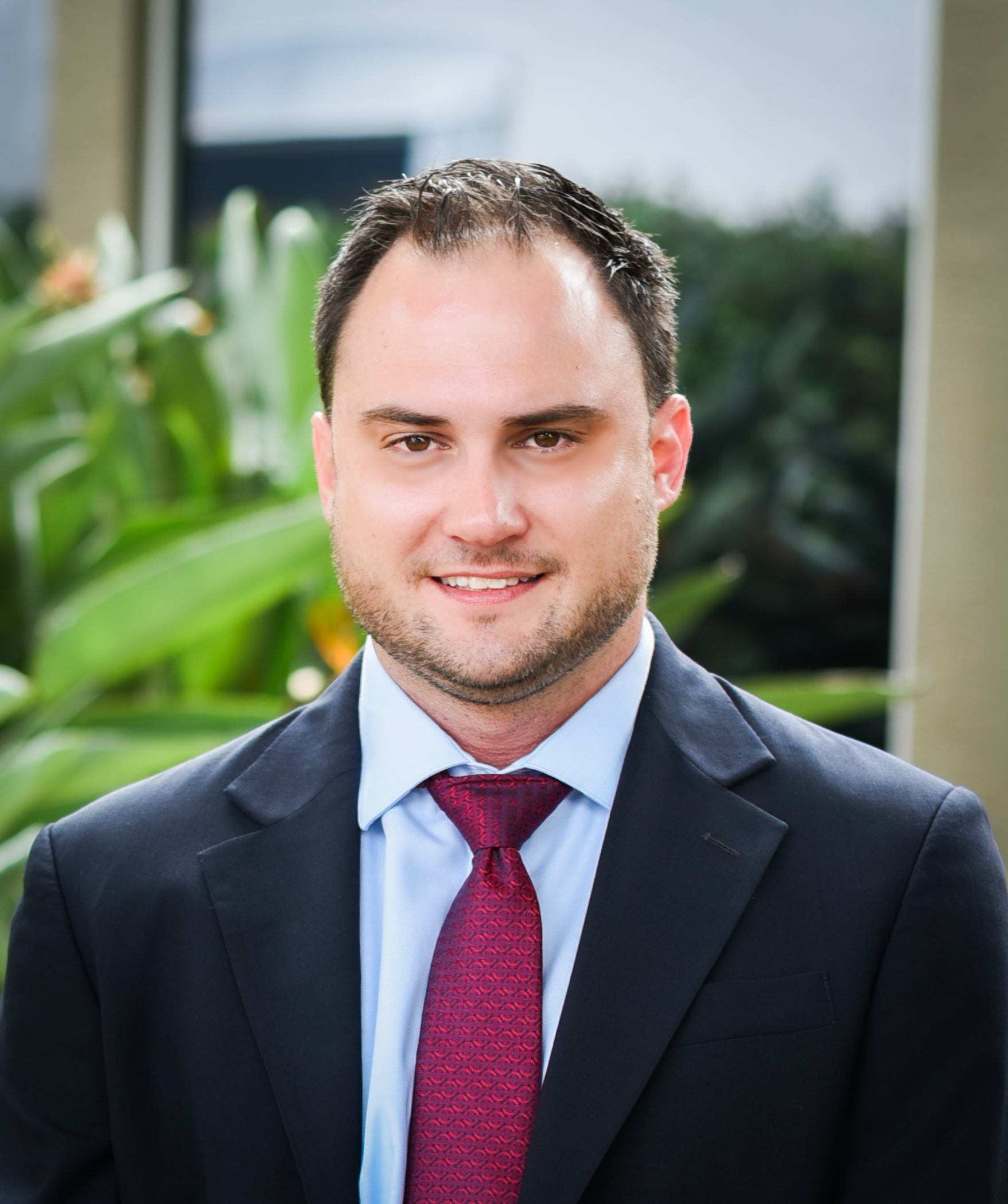 Hampton Law Man in a dark suit jacket, light blue dress shirt, and red patterned tie standing outdoors in front of green plants, smiling slightly.