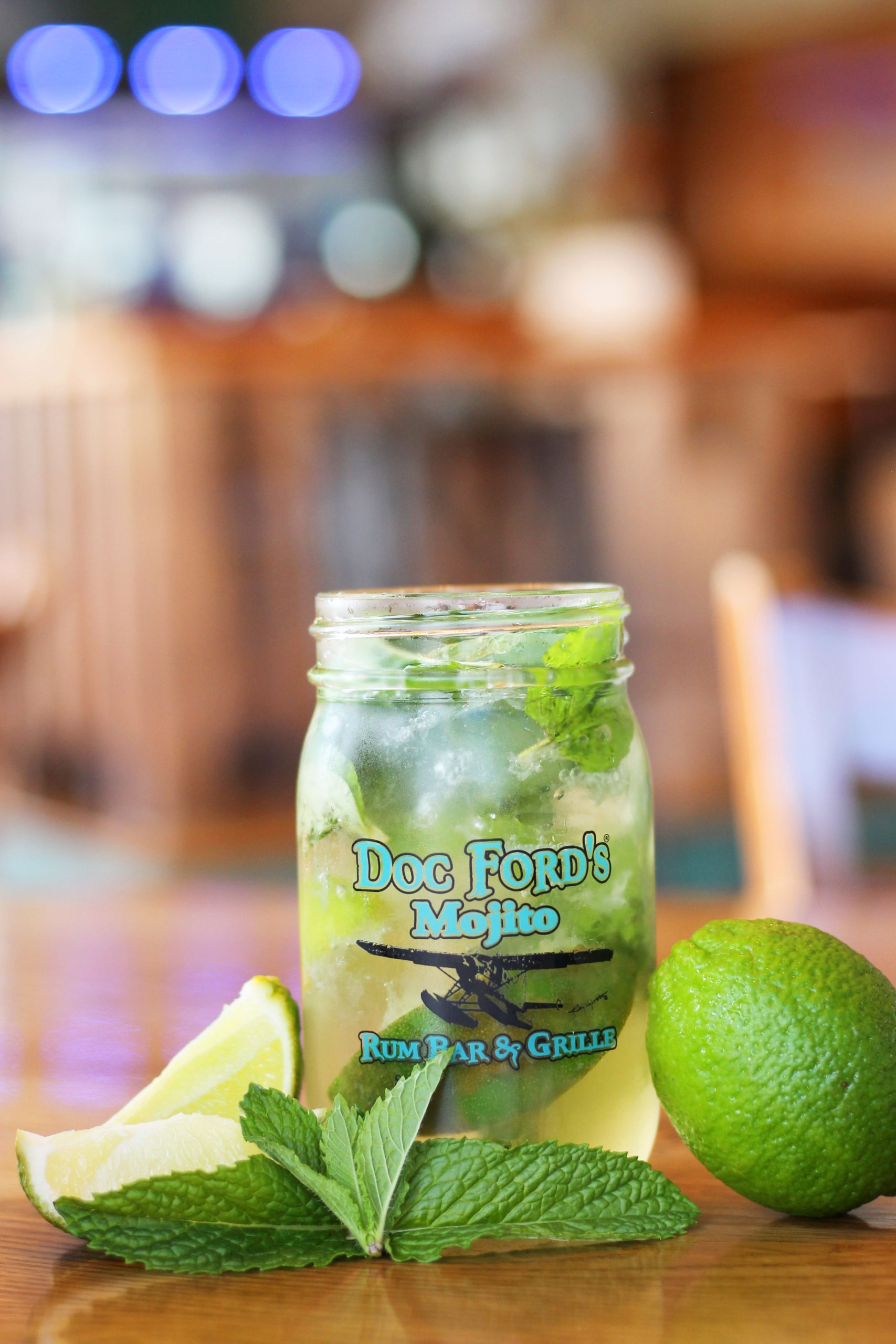 Doc-Fords-Rum-Ba-Grille-National-Mojito-Day-scaled-1 Mojito cocktail in Doc Ford's branded mason jar with fresh lime wedges and mint leaves on wooden bar counter.