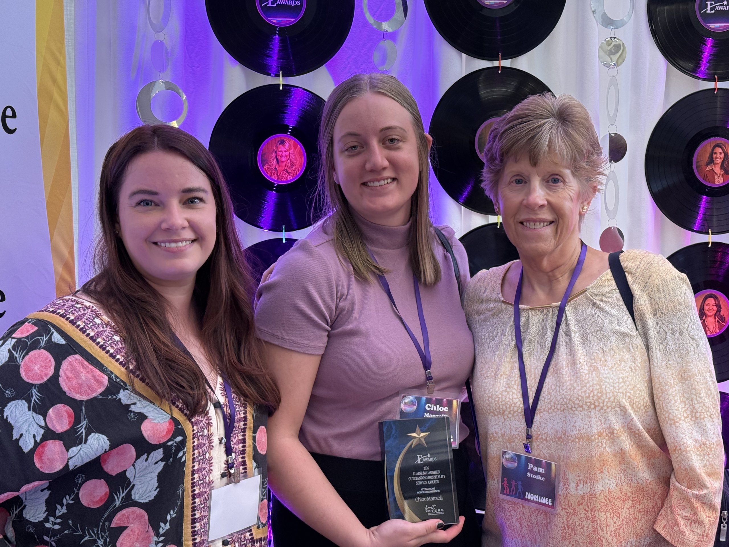 amber-chloe-pam-e-awards-2026-scaled-1 Three women smile with an award trophy at an event with vinyl record backdrop.